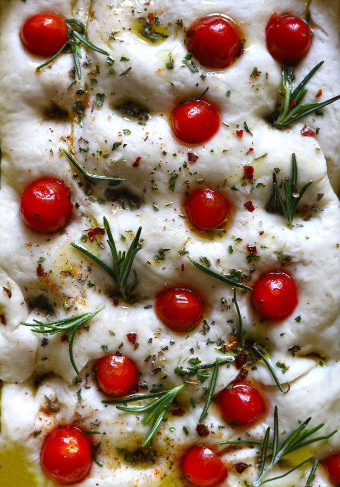 Aerial view of unbaked Focaccia bread topped with cherry tomatoes, rosemary, garlic and olive oil on baking tray