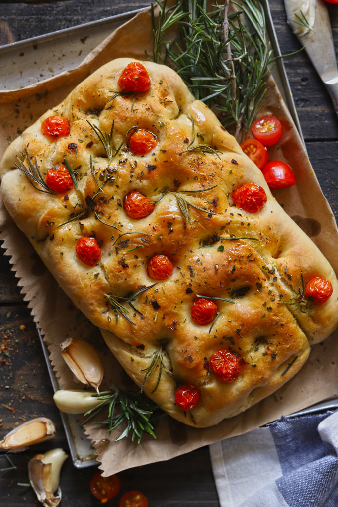 Focaccia bread topped with cherry tomatoes, rosemary and olive oil on cloth, with garlic beside