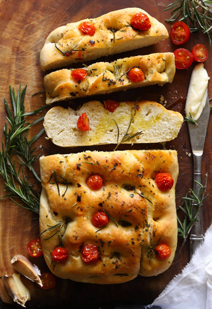 Slices of focaccia bread topped with cherry tomatoes, rosemary and olive oil on a wooden board, with garlic and rosemary beside.