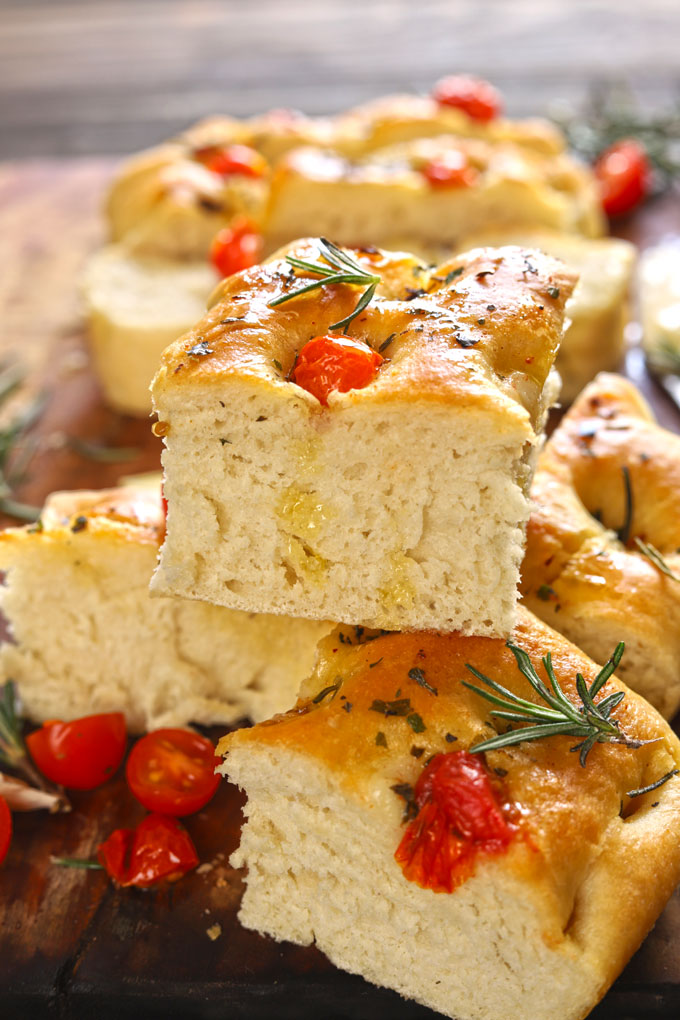 Focaccia bread topped with cherry tomatoes, rosemary and olive oil, cut into squares on wooden table