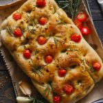 Focaccia bread topped with cherry tomatoes, rosemary and olive oil on cloth, with garlic beside