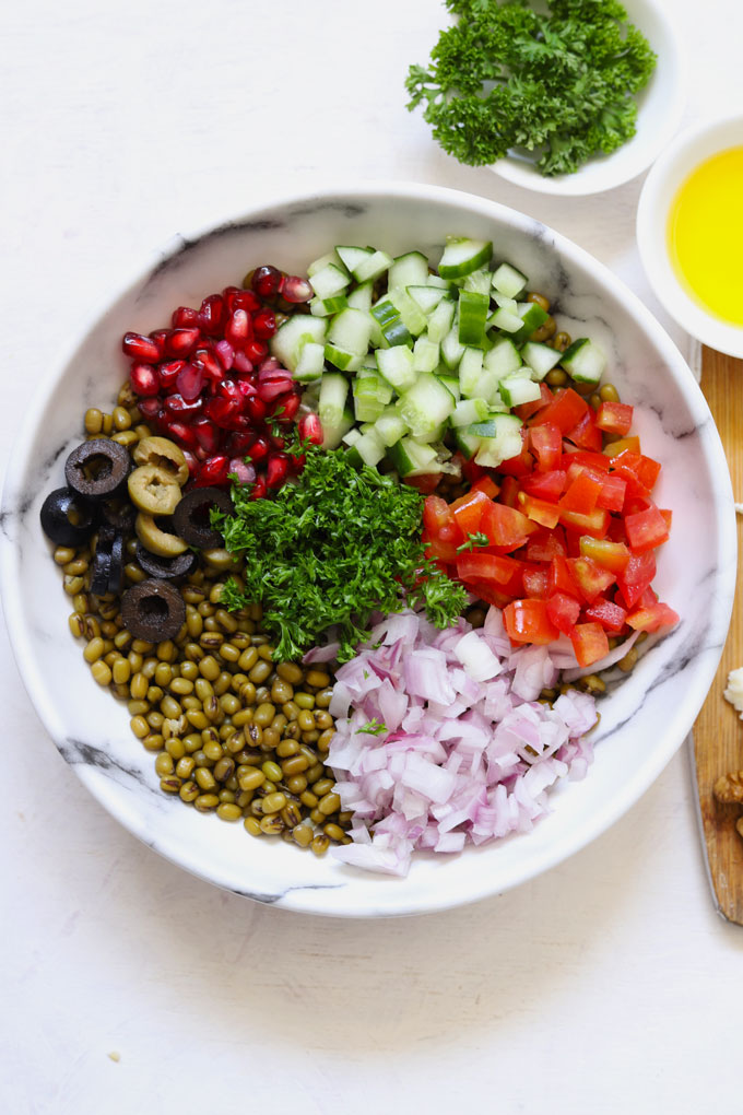 Aerial shot of boiled lentil, veggies, olives, parsley, pomegranate in a white bowl on a white background.