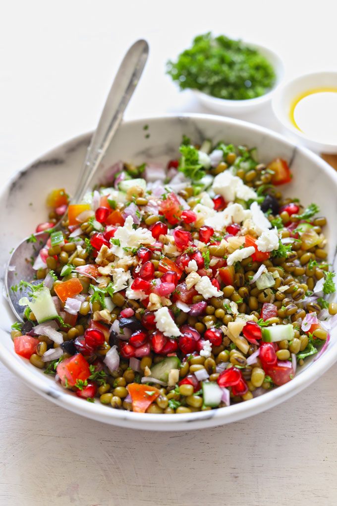 Side shot of lentil salad in a white bowl with serving spoons and parsley blur in the background.