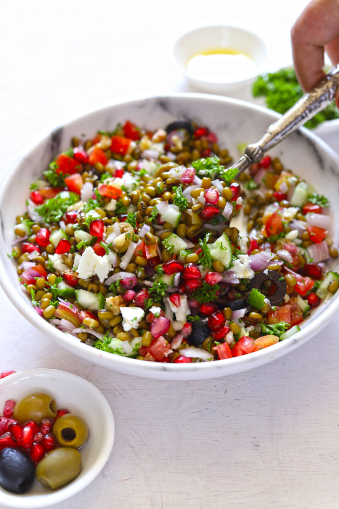 Side shot of lentil salad in a white bowl with serving spoons and parsley blur in the background.