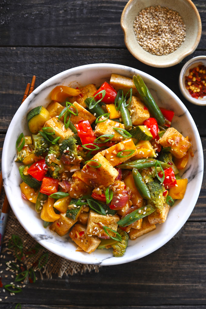 Aerial shot of white marble bowl with vegan tofu stir fry on dark wooden table, with napkin under bowl and chopsticks at the side. 2 small bowls of condiments in background.