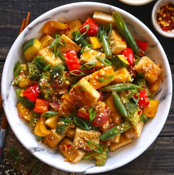 Aerial shot of white marble bowl with vegan tofu stir fry on dark wooden table, with napkin under bowl and chopsticks at the side. 2 small bowls of condiments in background.