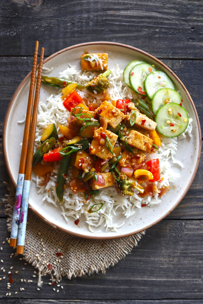 Cream stone plate with rice and vegan tofu stir fry on dark wooden table with chopsticks.