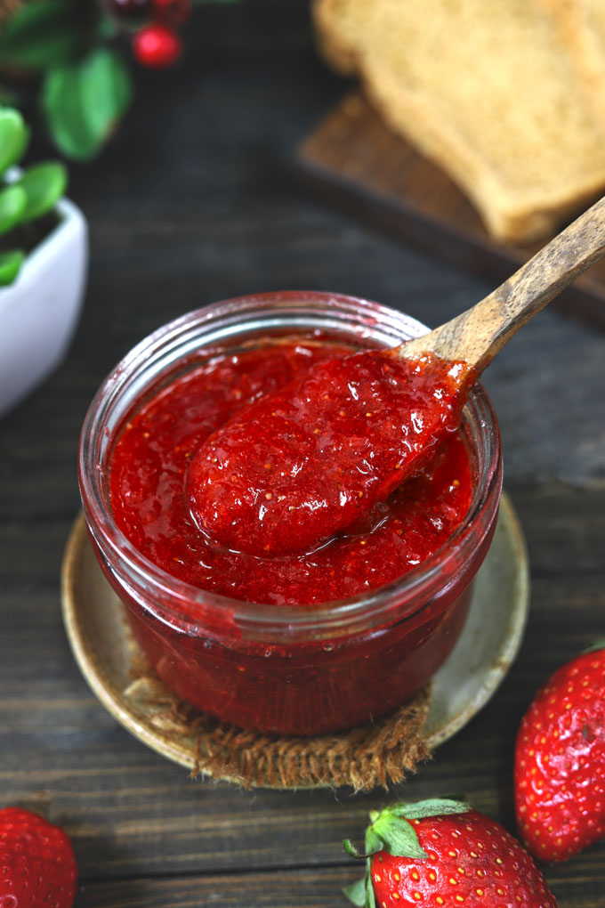 side shot of strawberry jam in a glass jar with a wooden spoon