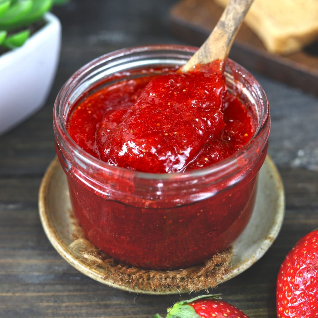 side shot of strawberry jam in a glass jar with a wooden spoon