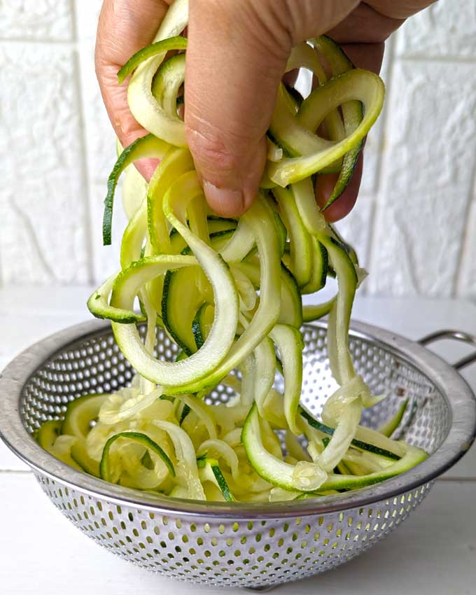 side shot of zoodles in a metal colander and woman hand picking the zoodles.